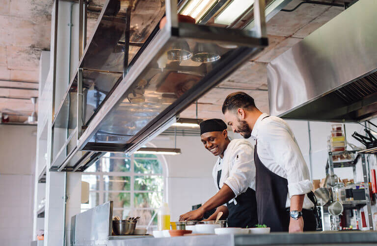 Two chefs in aprons talking and laughing in a brightly lit commercial kitchen, with one cooking while the other looks on.
