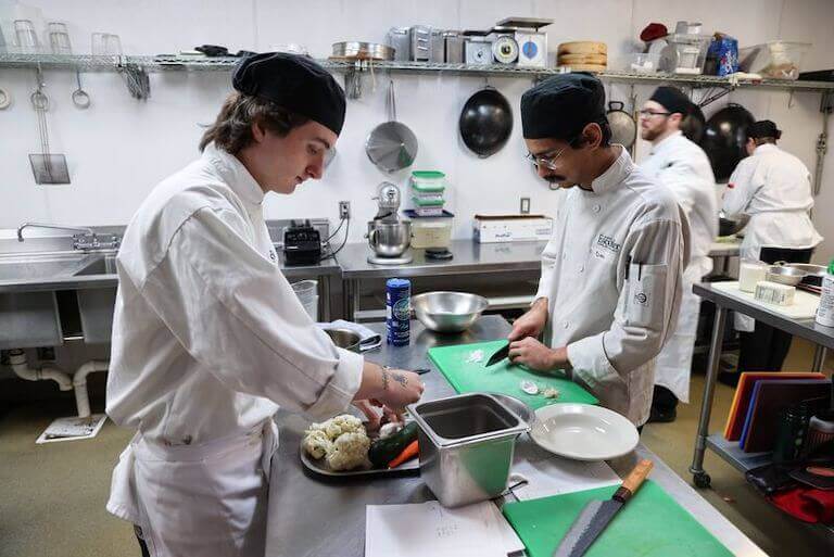 Two people in white culinary uniforms prepare ingredients in a kitchen at Escoffier’s Boulder campus.