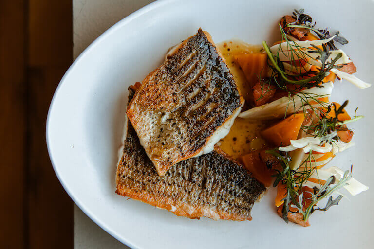 Two pieces of glazed roasted fish and a serving of roasted vegetables garnished with fresh herbs are beautifully plated on a white, oval dish.