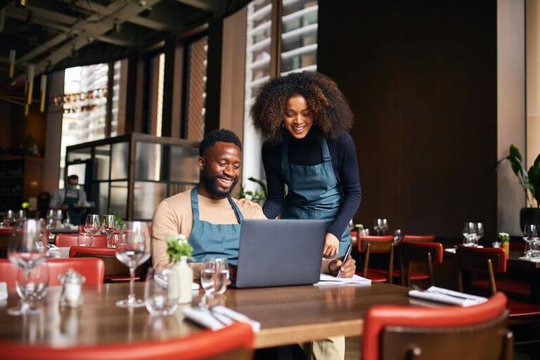 Two chefs wearing aprons reviewing information on a laptop in a restaurant.