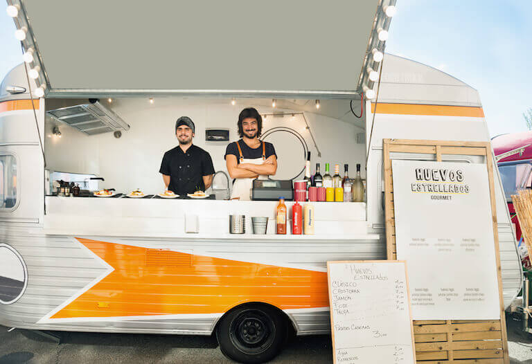 Two people stand inside of a food truck and face the camera, smiling. The food truck is called “Huevos Estrellados.”