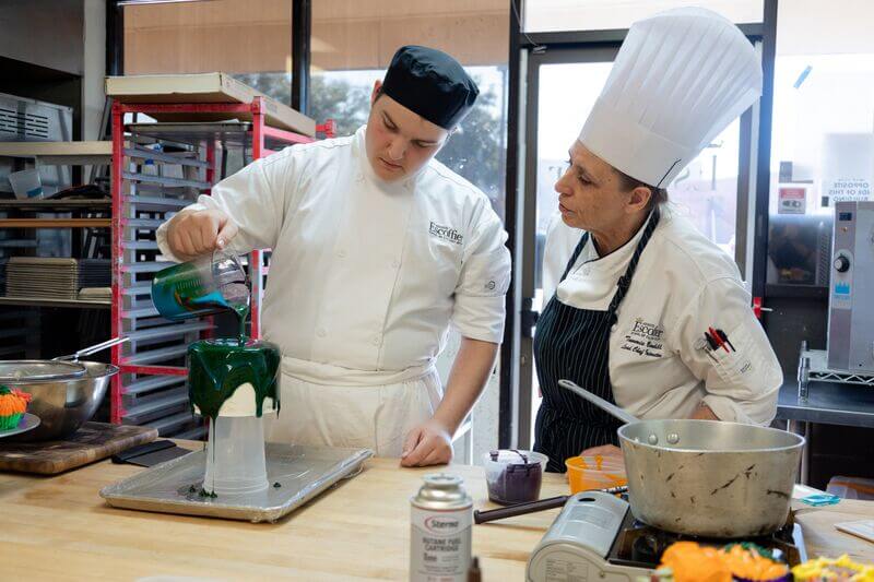Culinary student pours green glaze over a cake while a chef instructor observes and provides guidance in a professional kitchen classroom