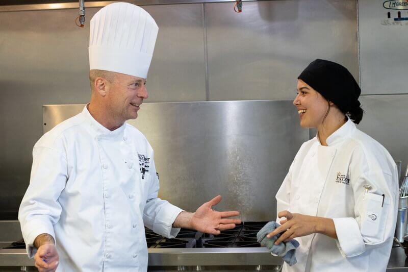 A chef instructor in a tall white hat and white Escoffier uniform smiles and gestures while speaking with a culinary student wearing a black cap and white jacket, both standing in a professional kitchen.