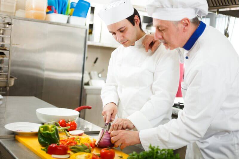 Two chefs in a commercial kitchen. One is chopping vegetables and another has a hand on their shoulder and is offering guidance.