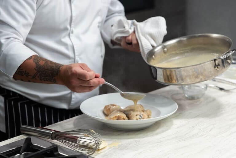A chef spoons sauce over chicken fricasee in a shallow white bowl, holding a steaming pan in one hand while a whisk rests on the countertop nearby.