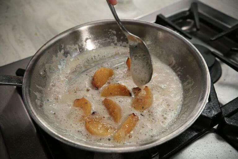 A chef’s hand sautés peach slices in butter in a stainless steel pan.