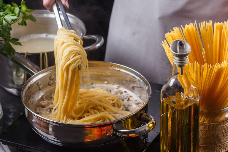 Close-up of spaghetti being added to a creamy sauce in a pan, with a pot of boiling water, a bottle of olive oil, and uncooked pasta in the background.