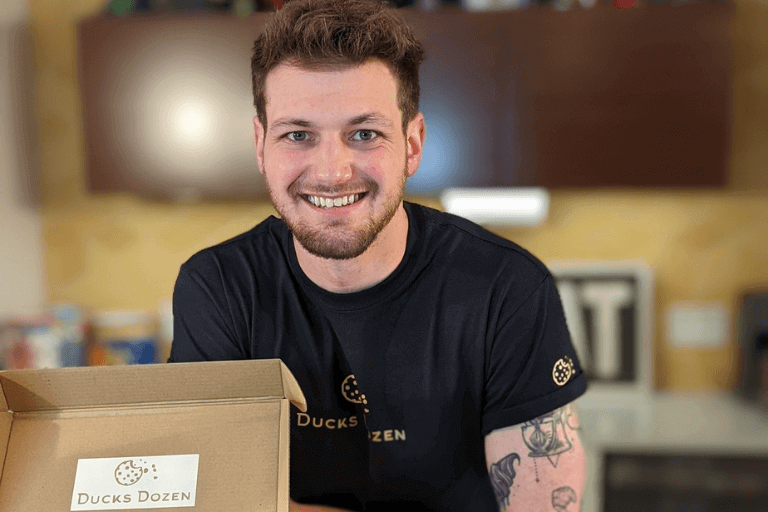 Wes Duckworth, Auguste Escoffier School of Culinary Arts graduate, smiling in black Ducks Dozen branded shirt while holding cookie delivery box in commercial kitchen.