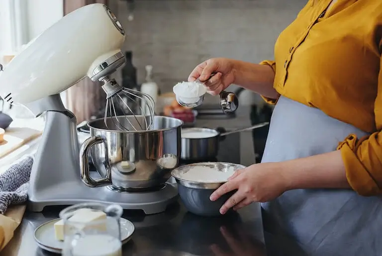 A baker in a yellow shirt measuring flour into a stand mixer bowl.