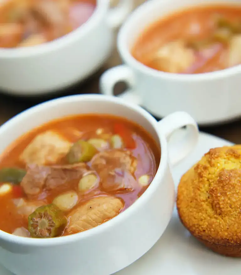 A bowl of burgoo, a traditional Kentucky stew, filled with meat, okra, corn, and vegetables, served alongside a golden cornbread muffin.