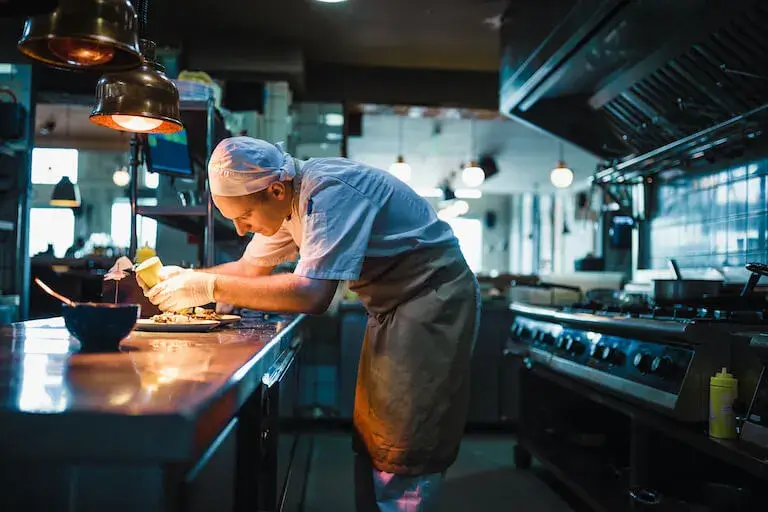 A chef leaning over the pass, plating food under warm lights in a restaurant kitchen.