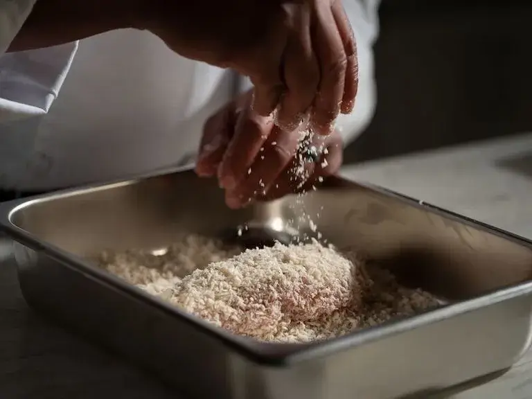 A chef's hands sprinkling bread crumbs over a piece of meat in a metal tray.