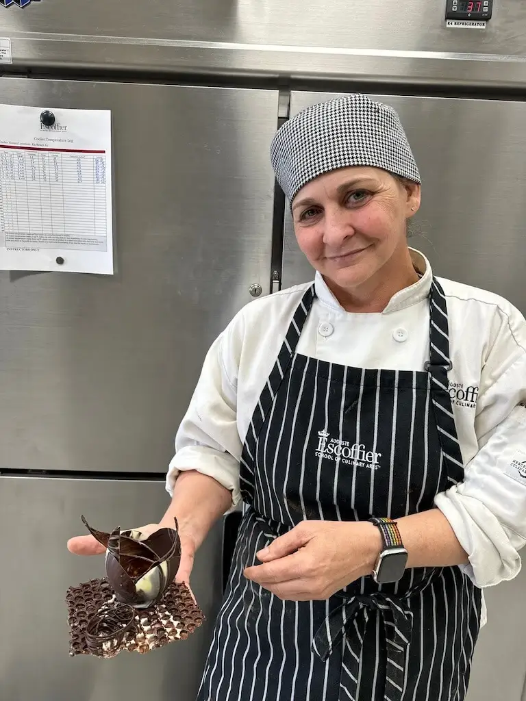 A culinary student in white chefs coat and striped apron holds an intricate chocolate dessert in a professional kitchen.