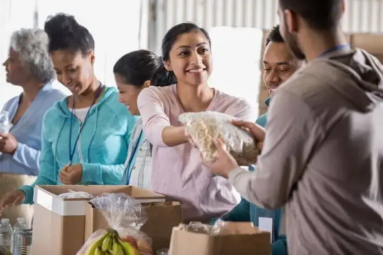 A line of people standing behind boxes with one person smiling and handing a bag of food to another.