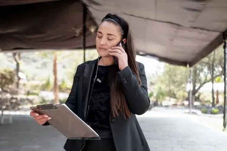 A person dressed in a suit speaking on the phone and looking at a clipboard while at an outdoor venue.