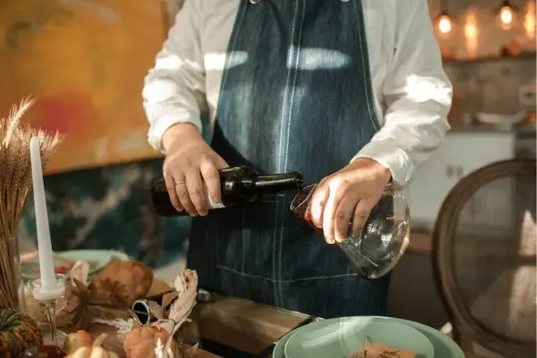 A person in a denim apron pours red wine into a decanter at a warmly lit table set for a meal.