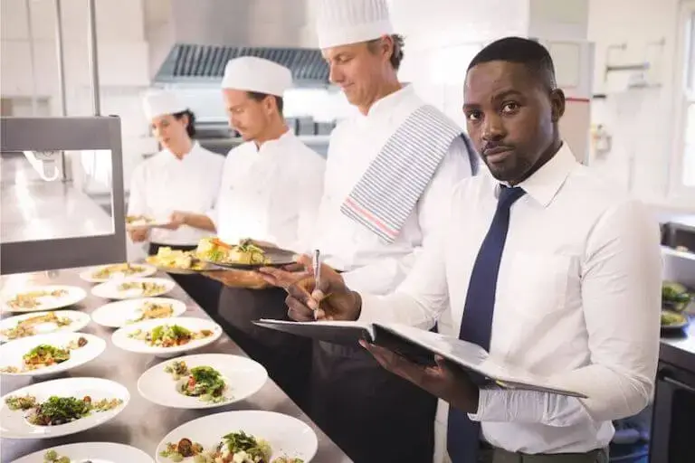 A person in a dress shirt and tie takes notes in a professional kitchen while three people in chefs coats plate dishes.