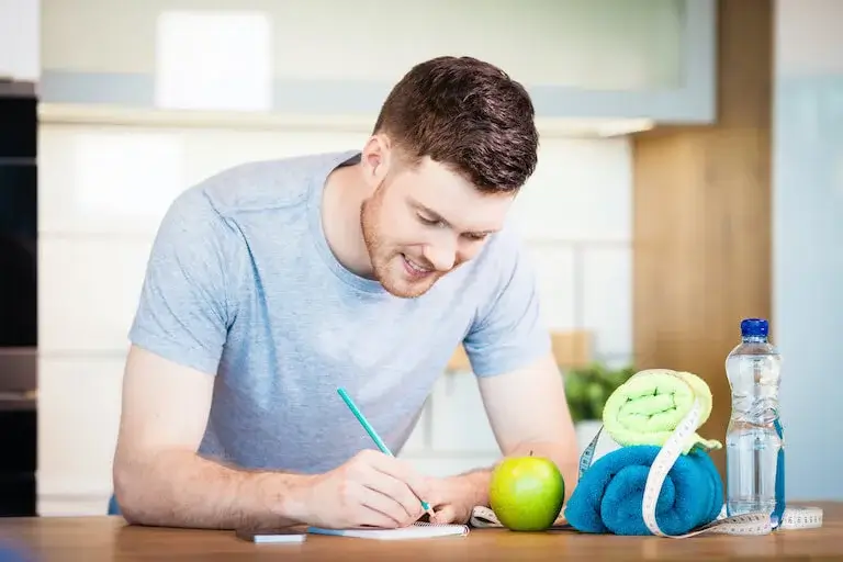 A person leans against a counter writing in a notebook beside an apple, bottle of water, towels, and other wellness items.