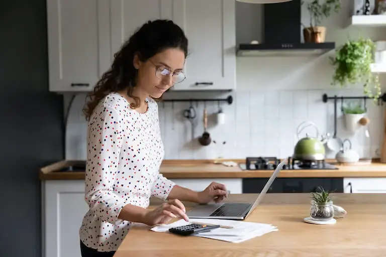 A person stands at their kitchen counter before an open laptop, a calculator, and a stack of papers.