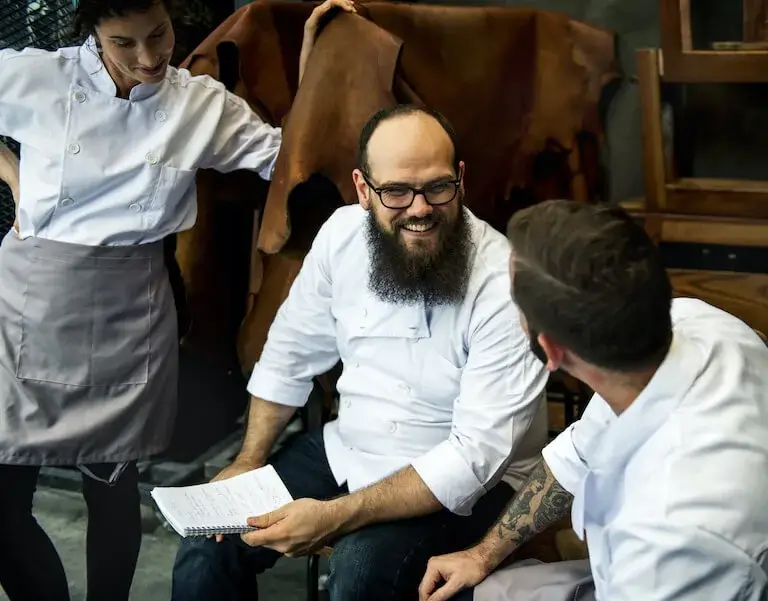 A smiling person with a beard and wearing a chef’s uniform holds a notebook while chatting with two other people also wearing chef’s uniforms.