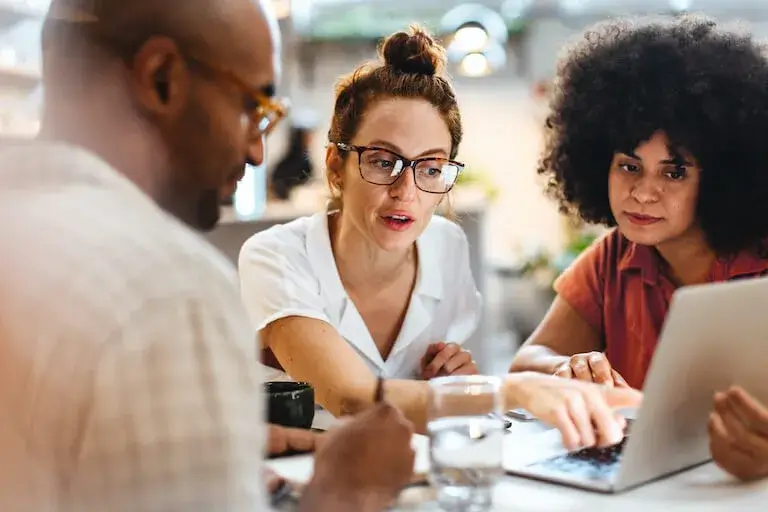 A trio of people sit at a table and look at a laptop, while the person in the middle points to the screen.