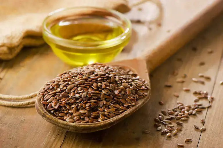 A wooden spoon filled with flax seeds next to a small bowl of oil on a rustic table.