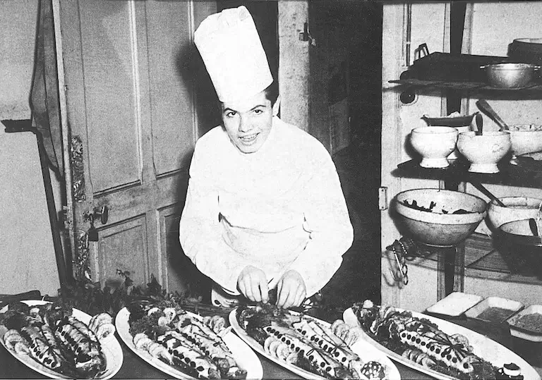 An early black-and-white photo of a young chef Jacques Pépin in white chef apron and chef's hat, working in a professional kitchen.