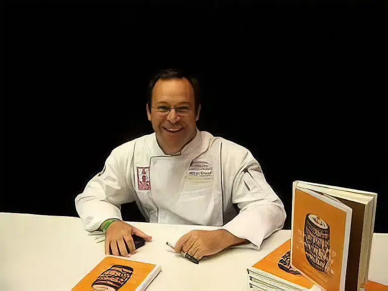 Chef Albert Schmid, wearing a white chef’s coat at a table, signing copies of his cookbook, with stacks of his book displayed beside him.