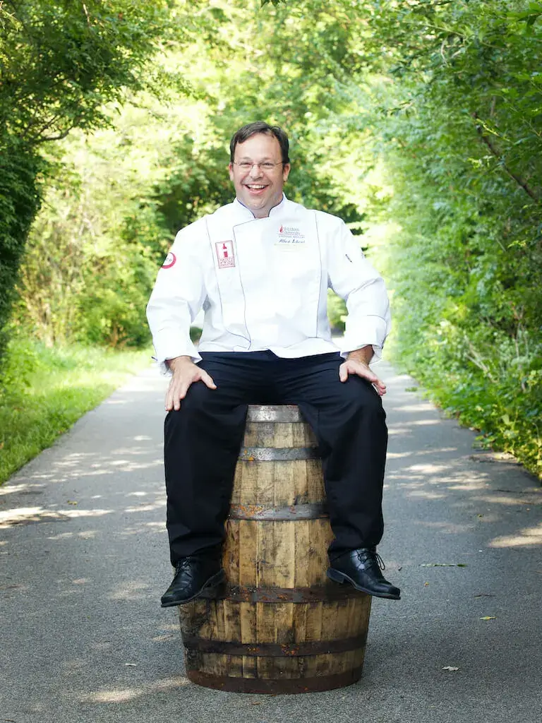 Chef Albert Schmid, wearing a white chef’s coat, sits on a wooden bourbon barrel along a tree-lined path, smiling.