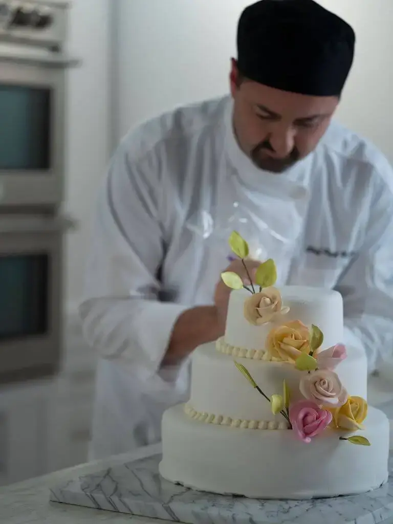 Chef decorating a three-tiered cake with frosting beads around each layer and gumpaste yellow and pink flowers winding up the tiers.
