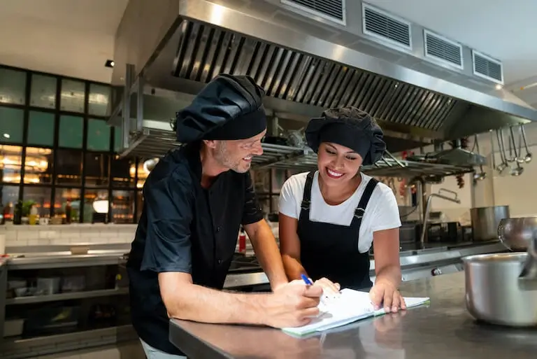Chef in black uniform and hat reviewing notes with a smiling chef in a commercial kitchen.