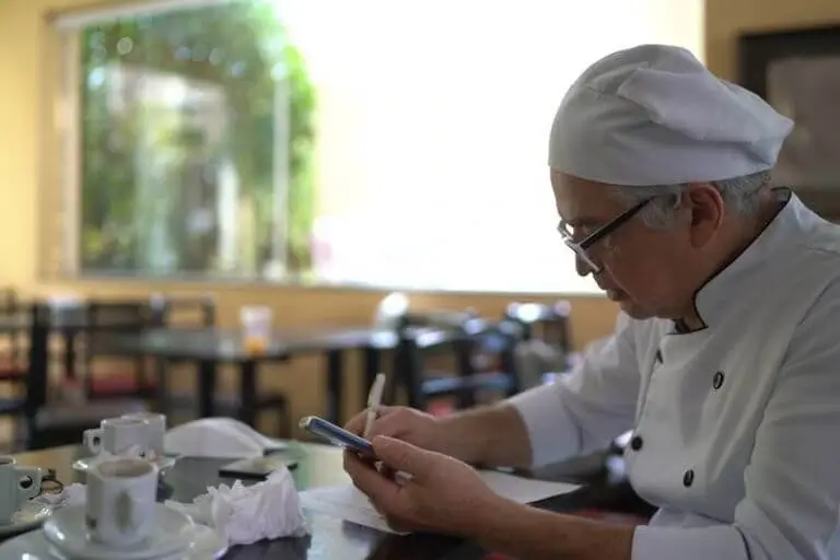 Chef in uniform sitting at a table reviewing financial notes and costs with a tablet.