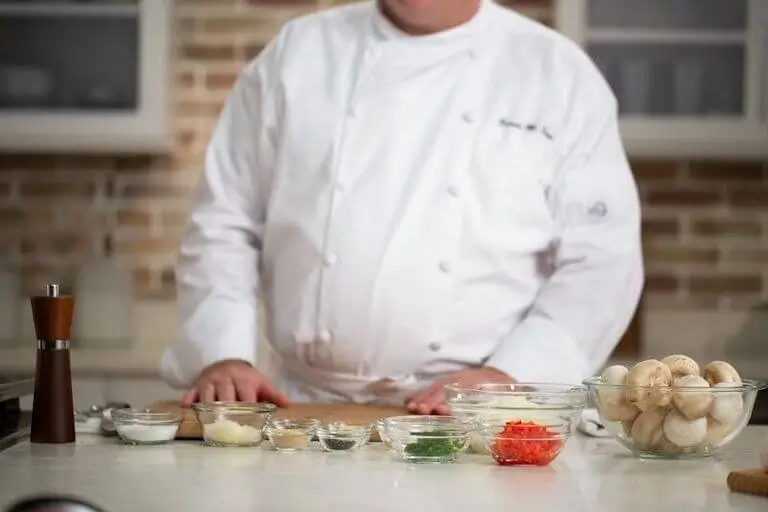Chef in uniform standing at a kitchen counter with organized mise en place, including mushrooms, spices, and prep bowls.