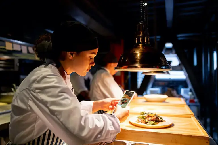 Chef taking a photo of plated food under a heat lamp in a restaurant kitchen.