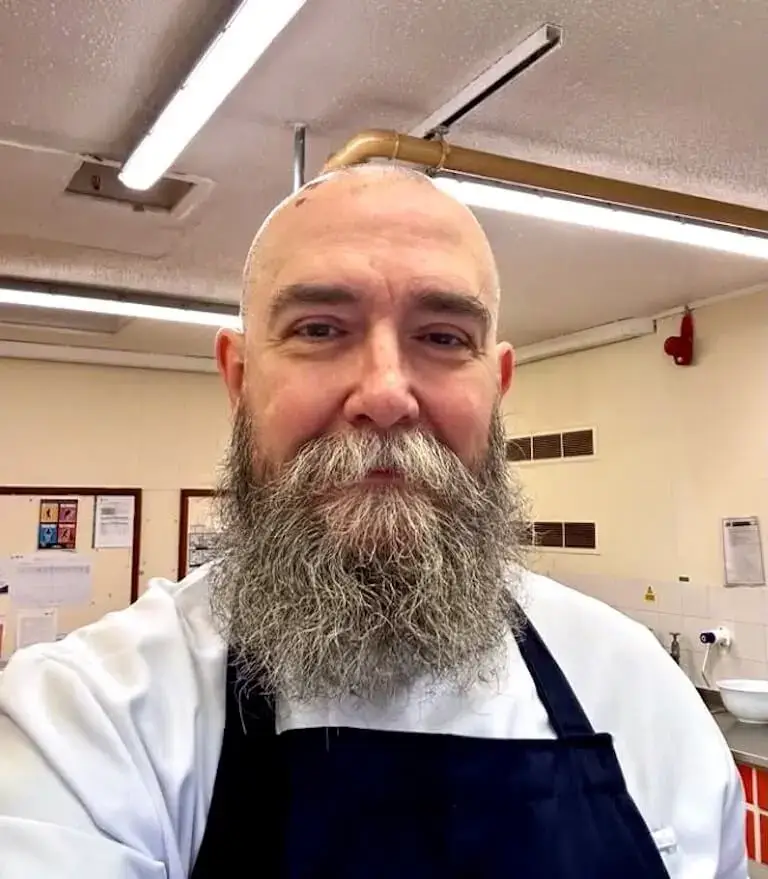 Chef Marc Mole wearing a white chef coat and dark apron, smiling in a professional kitchen.