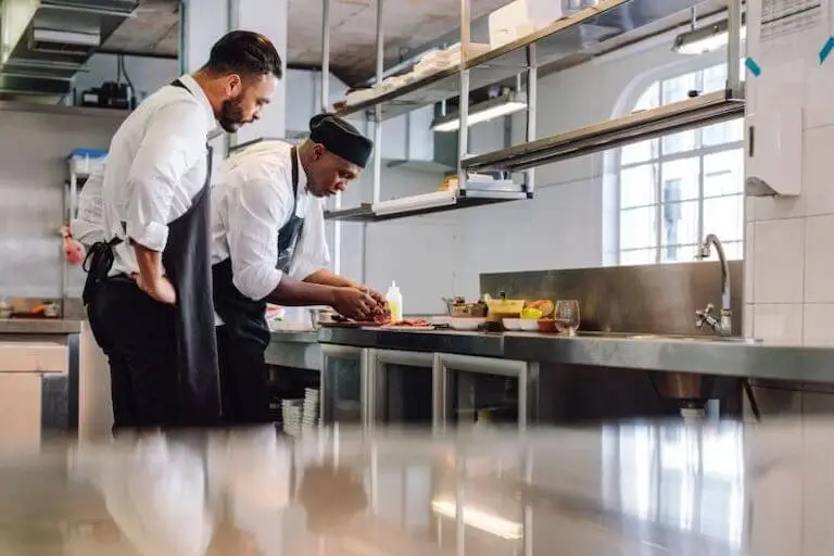 Chef mentoring a cook while preparing food together in a commercial kitchen.