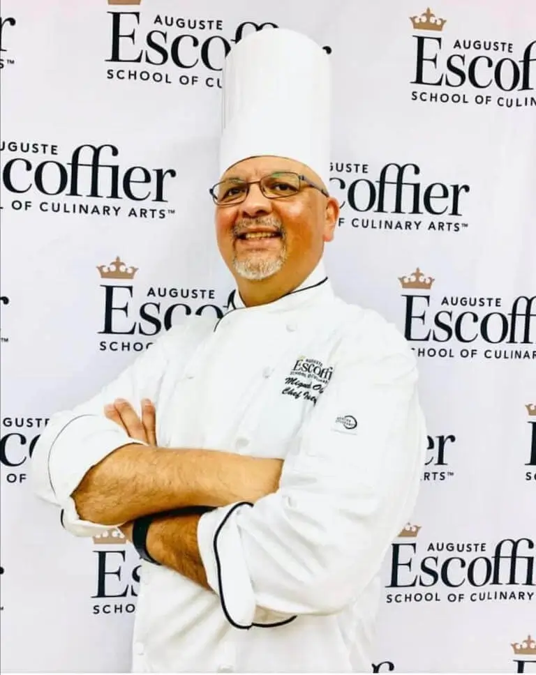 Chef Miguel Olmedo stands confidently in front of an Auguste Escoffier School of Culinary Arts backdrop, arms crossed and smiling.