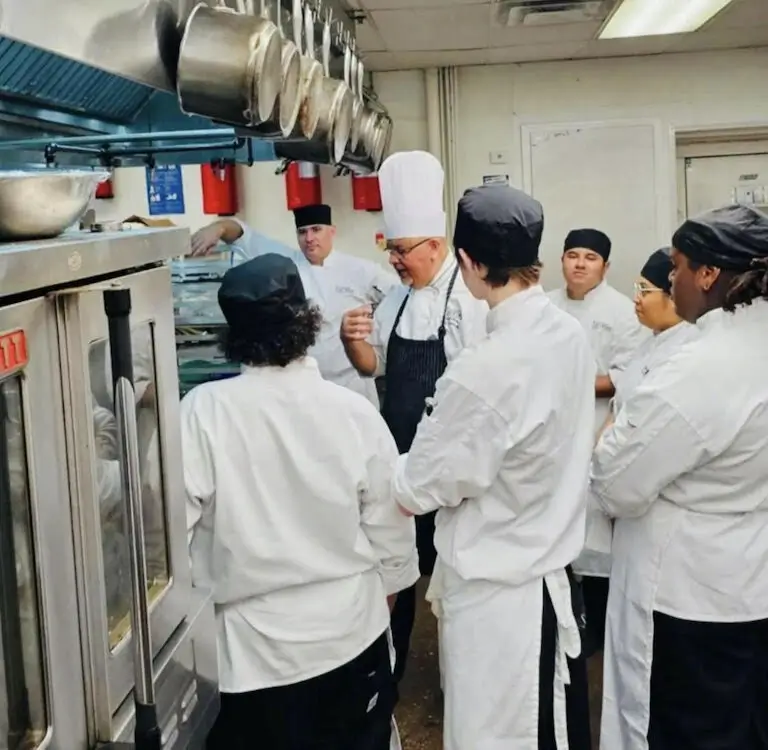 Chef Miguel Olmedo teaches a group of culinary students in a professional kitchen, gesturing as he explains a technique.