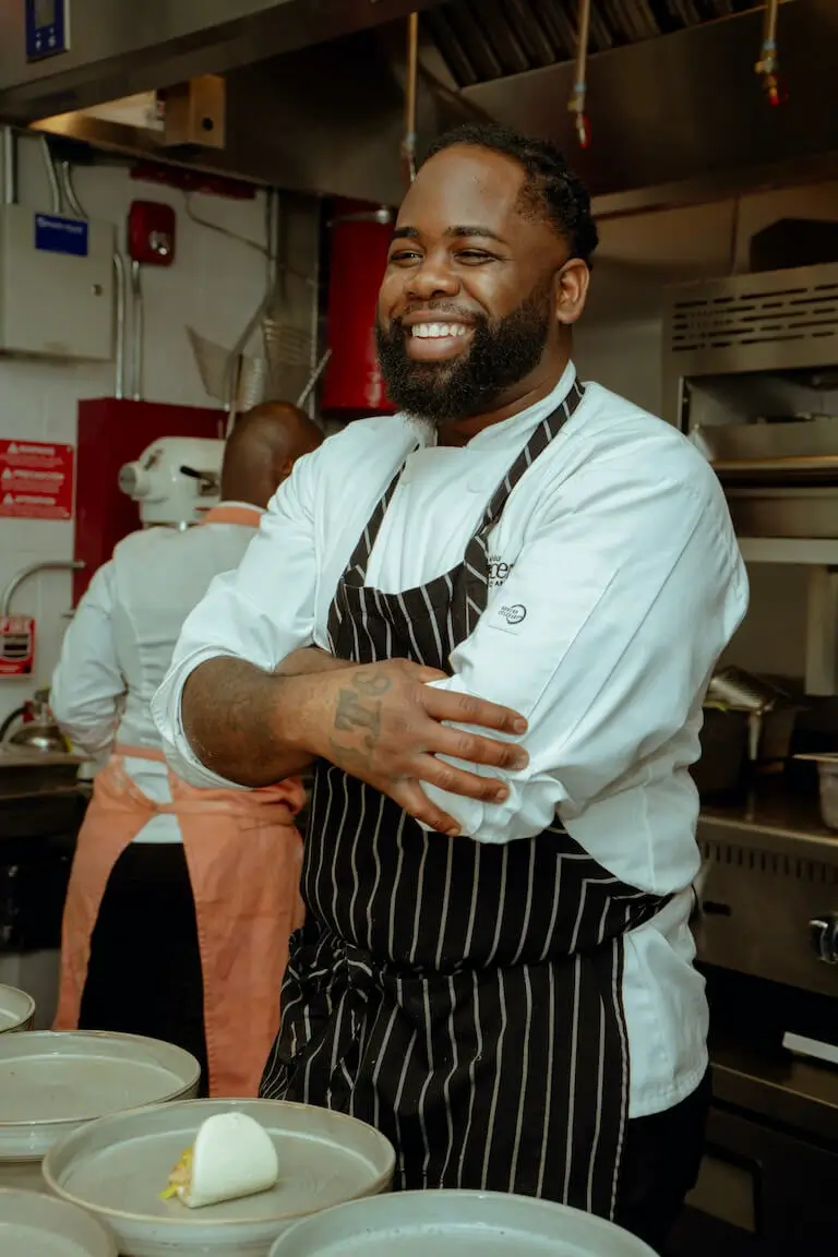 Chef Rodney Smith stands smiling with arms crossed in a commercial kitchen, wearing a white chef’s coat and a black pinstriped apron.