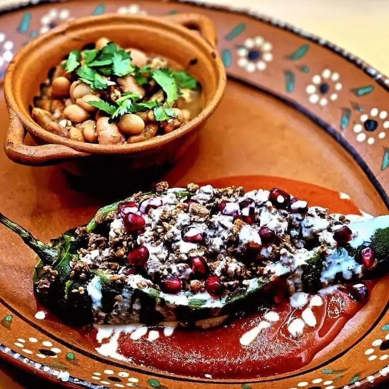 Close-up image of a chile relleno with chipotle sauce, crema, and a bowl of pinto beans on a hand-painted plate.