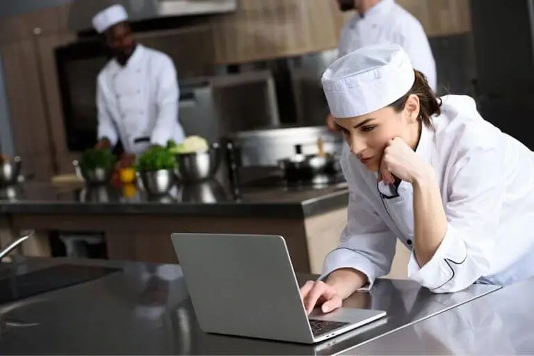 Culinary student in chef uniform studying on a laptop in a professional kitchen.