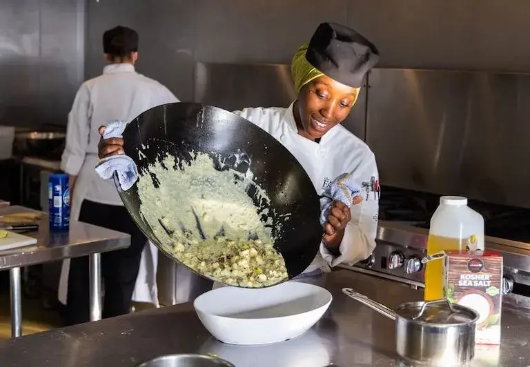 Culinary student pouring a creamy vegetable mixture from a wok into a serving dish during a cooking class.