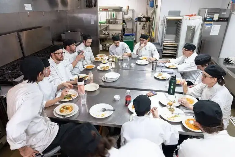 Culinary students in white chef coats gathered around a table, eating and talking.