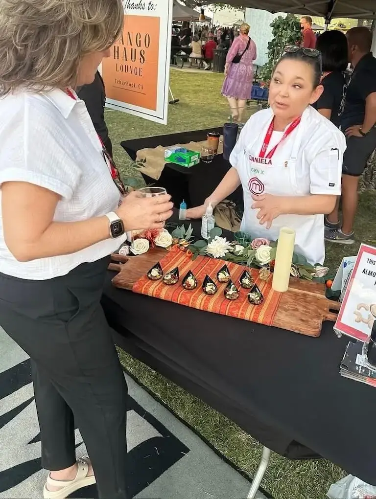 Daniela Peregrina, wearing a white MasterChef jacket, talks with an attendee at the Cheers to Charity event in Tehachapi, California. On the table in front of her is a wooden board lined with small black spoons filled with appetizers inspired by her MasterChef dish, Chiles en Nogada.