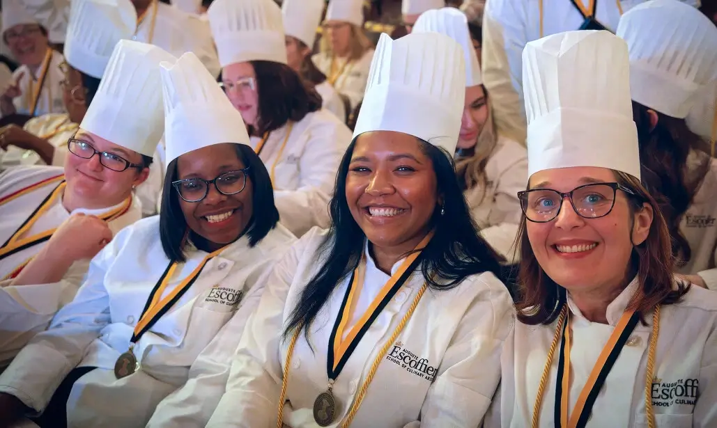 Group of Escoffier culinary graduates smiling in white chef coats and hats during commencement ceremony