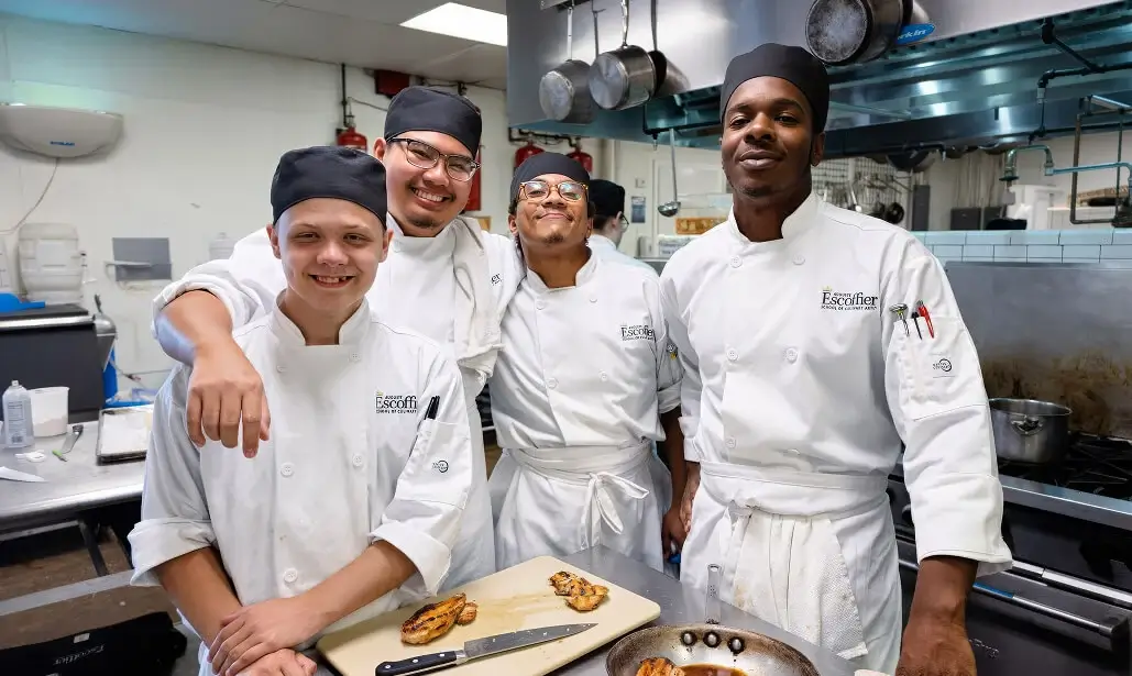 Four Escoffier culinary students smile together in a commercial kitchen, wearing white chef coats and black hats