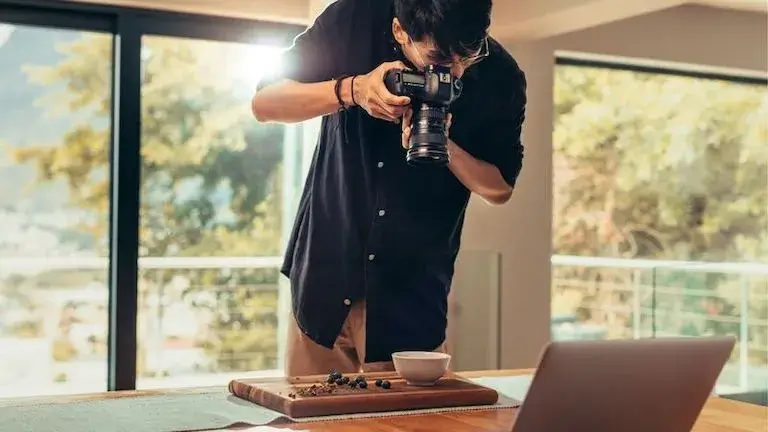 Food photographer taking overhead shot of plated dish with blueberries on wooden cutting board, with laptop visible in background.