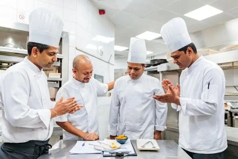 Four chefs in a commercial kitchen reviewing a recipe together, with two chefs applauding.