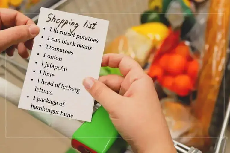 Hands holding a printed grocery list over a shopping cart filled with groceries.