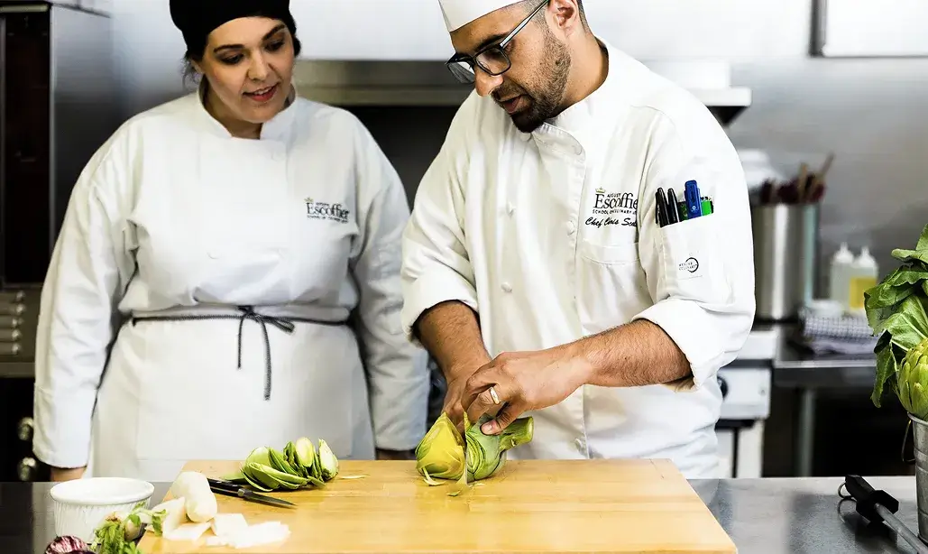 An Escoffier Chef Instructor slices an artichoke on a wooden board while a student observes attentively beside him in the kitchen